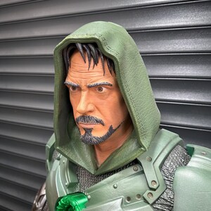 May include: A detailed bust of a character wearing a green hooded cloak and armor. The figure has a stern expression, a goatee, and dark hair. The armor features metallic accents and a glowing green emblem.