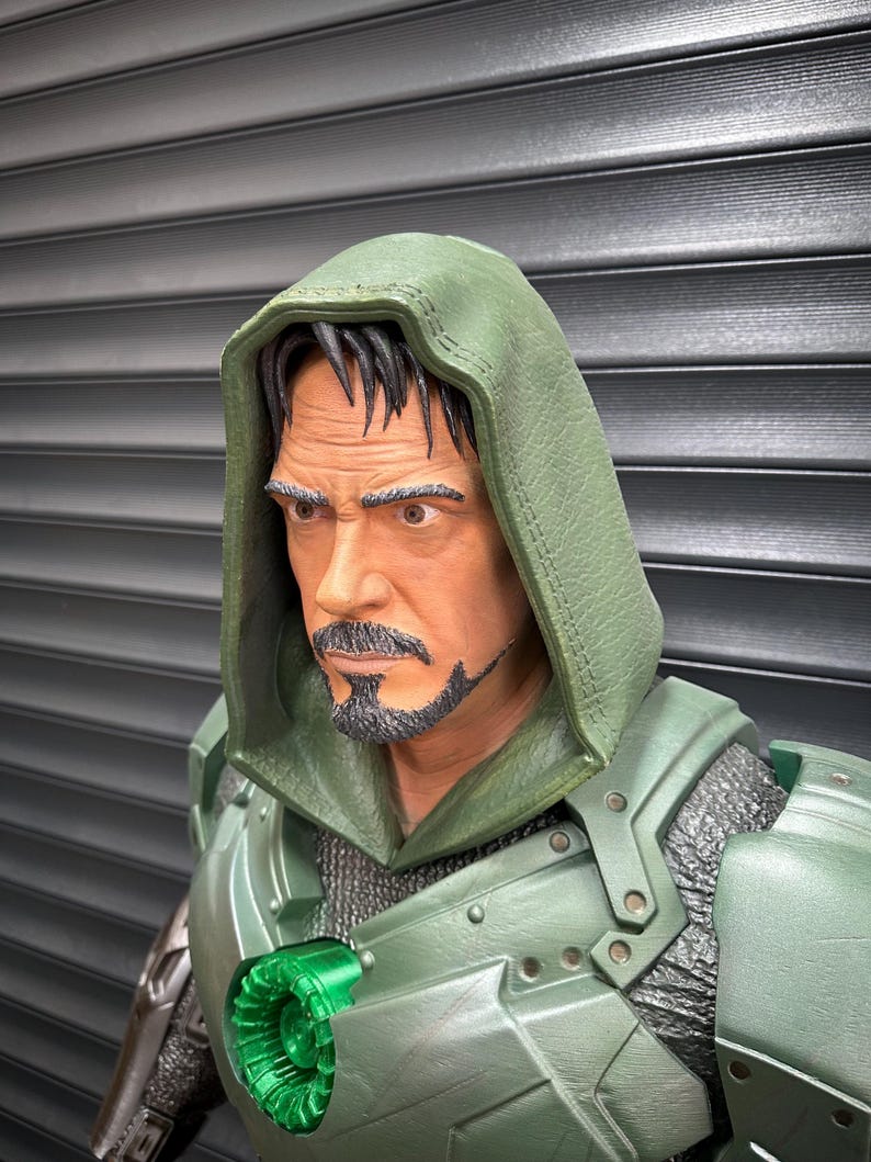 May include: A detailed bust of a character wearing a green hooded cloak and armor. The figure has a stern expression, a goatee, and dark hair. The armor features metallic accents and a glowing green emblem.