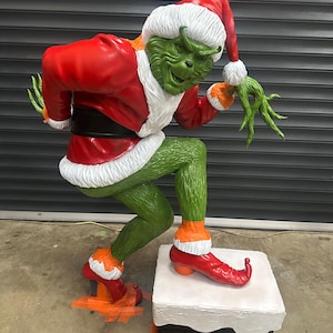 May include: A large, green Grinch statue in a Santa Claus outfit, complete with a red coat, hat, and boots. The statue is posed on a chimney, with a mischievous expression. The Grinch's outfit has white fur trim.