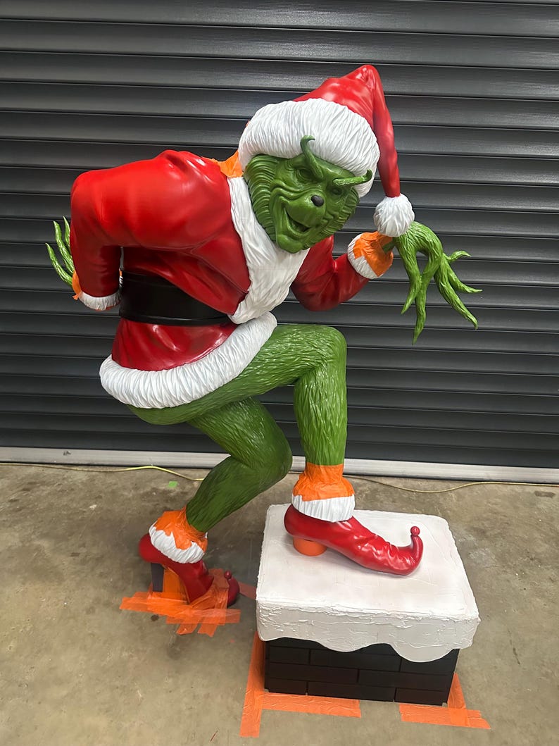 May include: A large, green Grinch statue in a Santa Claus outfit, complete with a red coat, hat, and boots. The statue is posed on a chimney, with a mischievous expression. The Grinch's outfit has white fur trim.