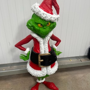 May include: A green character statue wearing a red Santa suit with white trim and a matching hat. The statue has a black belt with a gold buckle and red shoes. The character has a grumpy expression.