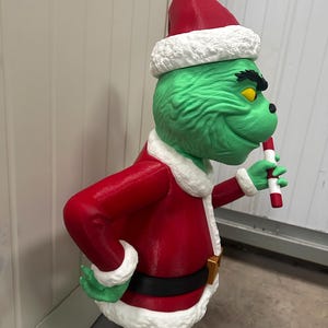 May include: A green character statue wearing a red Santa suit with white trim and a matching hat. The statue holds a candy cane and has a black belt with a gold buckle. The character has yellow eyes and a mischievous grin.