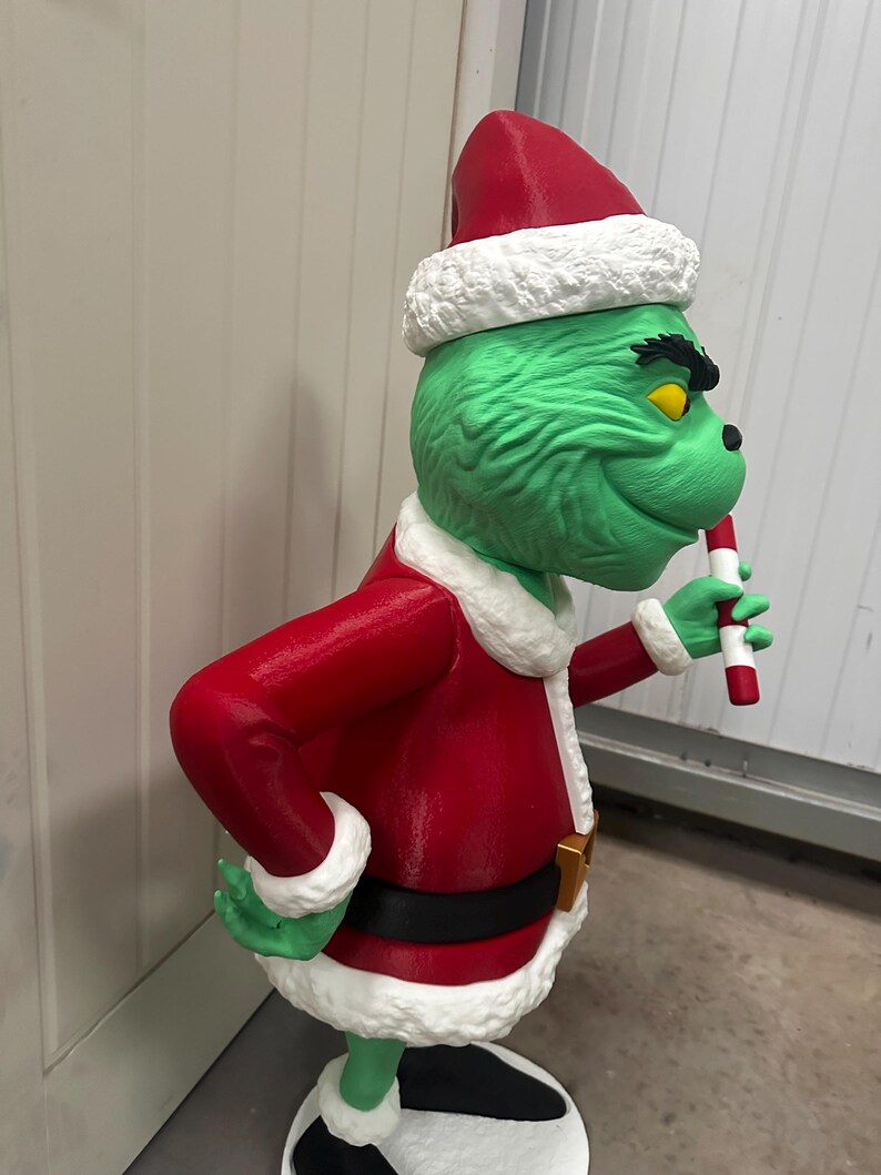 May include: A green character statue wearing a red Santa suit with white trim and a matching hat. The statue holds a candy cane and has a black belt with a gold buckle. The character has yellow eyes and a mischievous grin.