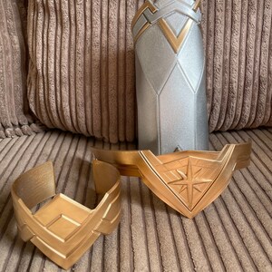 May include: Two gold-coloured Wonder Woman bracelets with a star design. The bracelets are on a brown corduroy surface.