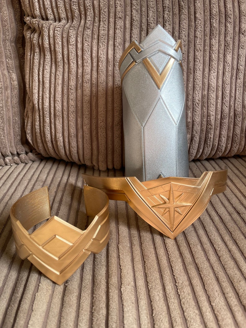 May include: Two gold-coloured Wonder Woman bracelets with a star design. The bracelets are on a brown corduroy surface.