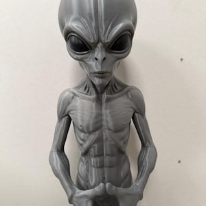 Pode incluir: Uma figura alien&iacute;gena cinza impressa em 3D com uma cabe&ccedil;a grande, lisa e alongada, com olhos pretos s&oacute;lidos em forma de am&ecirc;ndoa. A figura tem um torso esguio e musculoso com costelas e abdominais vis&iacute;veis, e bra&ccedil;os longos com as m&atilde;os juntas.