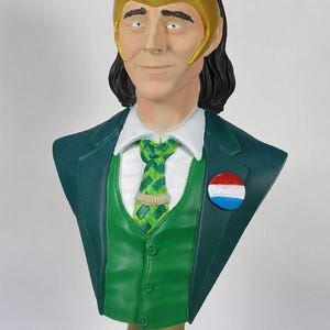 May include: A bust of a character with a green suit, vest, and tie. The bust has a gold-colored horned headpiece and a pin with red, white, and blue circles. The base is gold with the name "LOKI" in green.