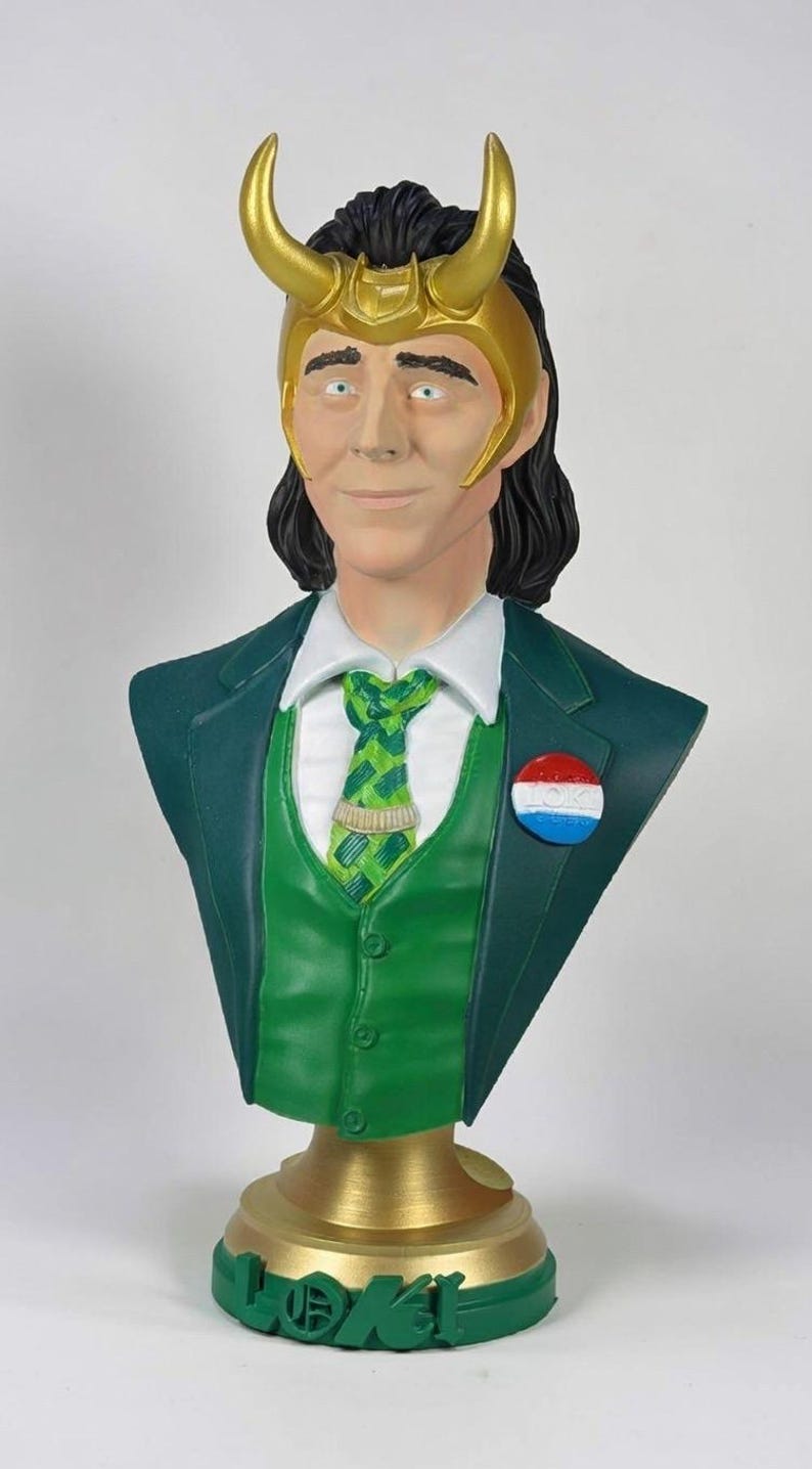 May include: A bust of a character with a green suit, vest, and tie. The bust has a gold-colored horned headpiece and a pin with red, white, and blue circles. The base is gold with the name "LOKI" in green.