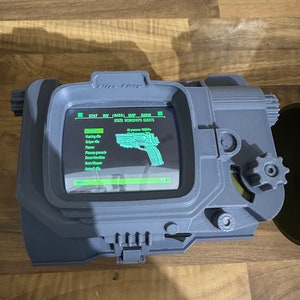 May include: A grey 3D printed replica of a Pip-Boy from the Fallout video game series. The screen displays a list of items, including a hunting rifle, sniper rifle, flamer, plasma grenade, brass knuckles, a 10mm pistol, and an assault rifle. The screen also displays the text "STATS WORKSHOPS QUESTS".
