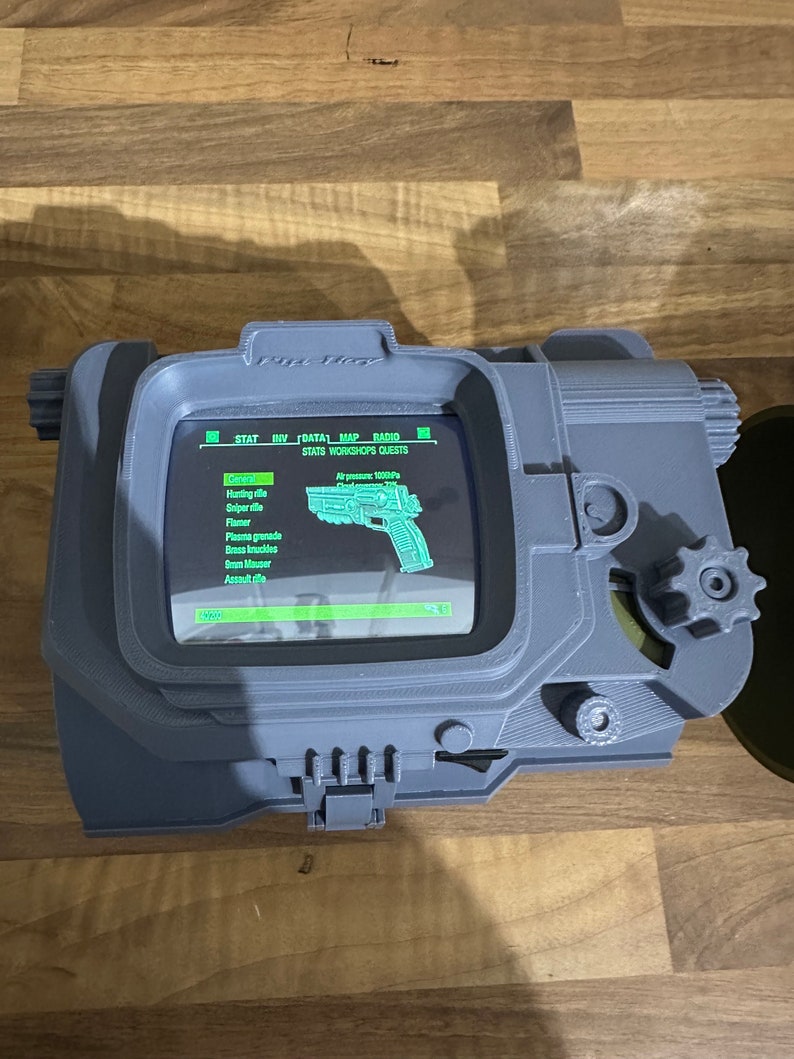 May include: A grey 3D printed replica of a Pip-Boy from the Fallout video game series. The screen displays a list of items, including a hunting rifle, sniper rifle, flamer, plasma grenade, brass knuckles, a 10mm pistol, and an assault rifle. The screen also displays the text "STATS WORKSHOPS QUESTS".