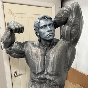 May include: A gray sculpture of a muscular man flexing his biceps. The statue has detailed facial features and is posed with one arm raised above the head. The sculpture is in a workshop setting.