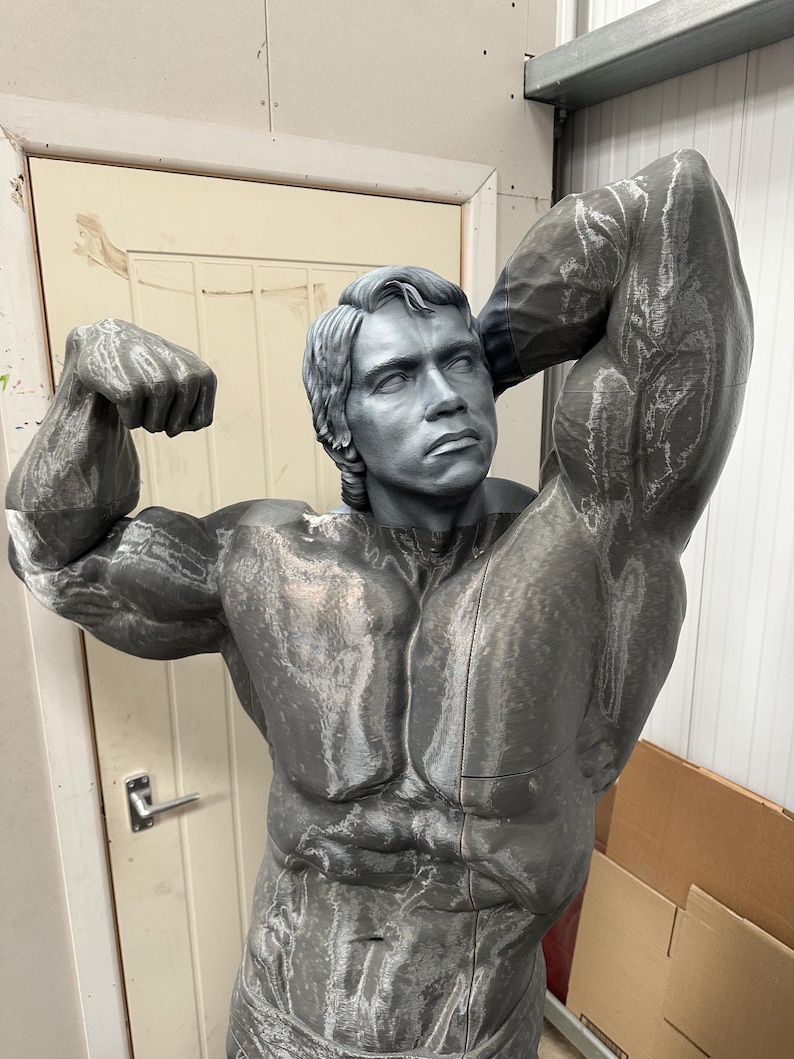 May include: A gray sculpture of a muscular man flexing his biceps. The statue has detailed facial features and is posed with one arm raised above the head. The sculpture is in a workshop setting.