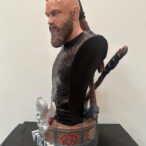 May include: A detailed bust of a Viking warrior with a beard and braided hair. The figure wears a black tunic and has a stern expression. Axes and a shield are visible behind the bust. The base features intricate designs and symbols.