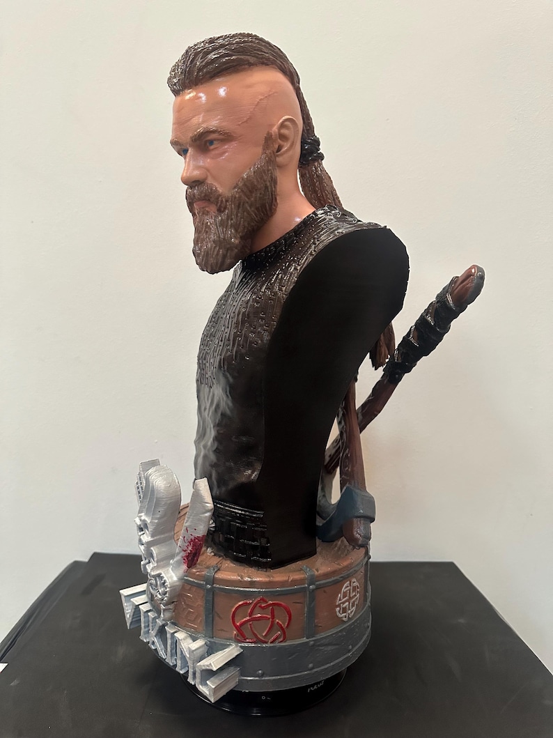 May include: A detailed bust of a Viking warrior with a beard and braided hair. The figure wears a black tunic and has a stern expression. Axes and a shield are visible behind the bust. The base features intricate designs and symbols.