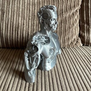May include: A silver-colored 3D printed bust of a robot with a detailed, exposed mechanical head and torso. The bust is posed with one arm extended and the other arm bent at the elbow.
