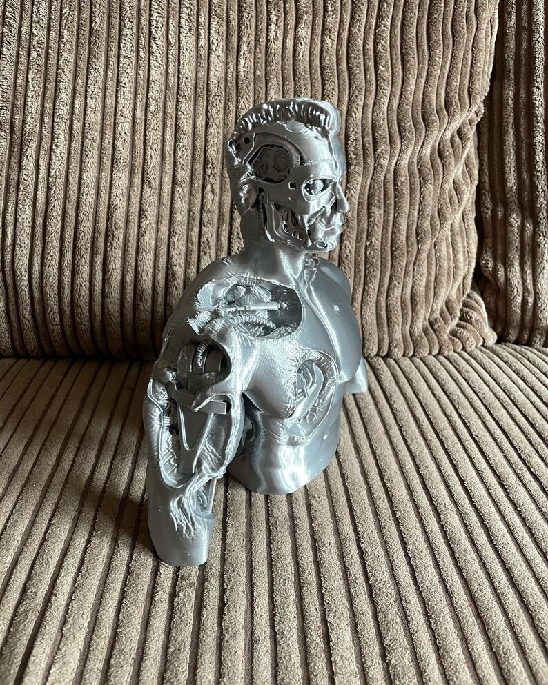 May include: A silver-colored 3D printed bust of a robot with a detailed, exposed mechanical head and torso. The bust is posed with one arm extended and the other arm bent at the elbow.