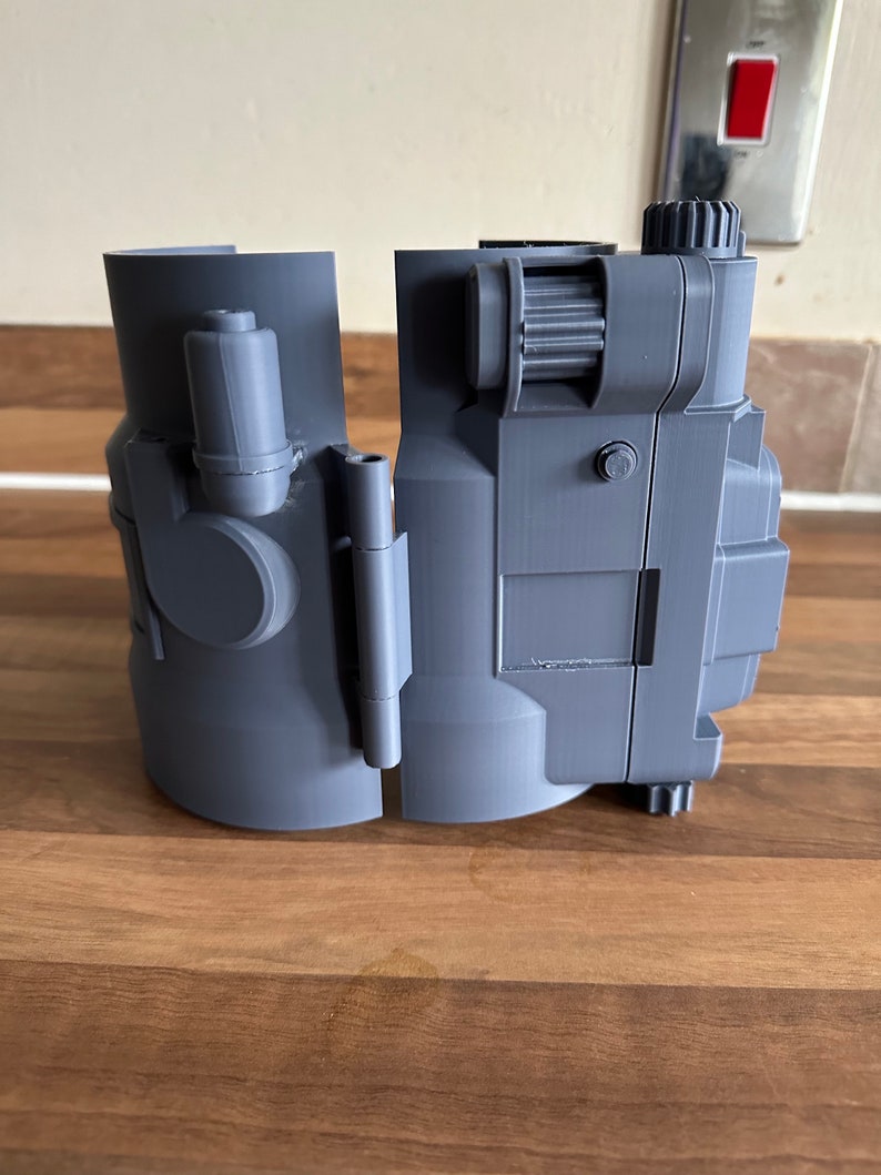 May include: A grey 3D printed model of a futuristic device with a gear on the side. The device is split into two pieces.