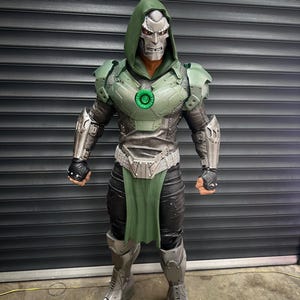 May include: A full-body shot of a detailed costume resembling a comic book character. The costume features a silver mask, a green hooded cloak, and metallic armor with green accents. The figure is standing in front of a gray, slatted background.