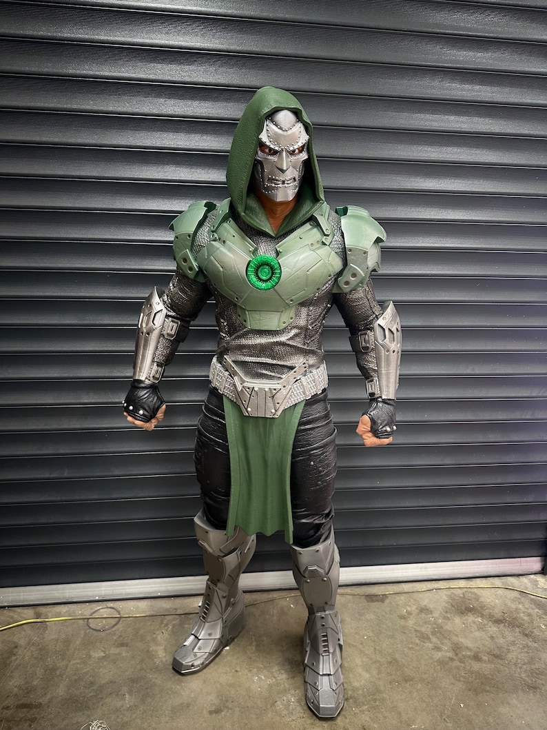 May include: A full-body shot of a detailed costume resembling a comic book character. The costume features a silver mask, a green hooded cloak, and metallic armor with green accents. The figure is standing in front of a gray, slatted background.