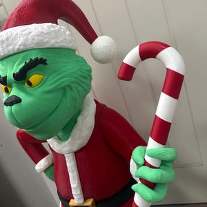 May include: A green character statue wearing a red Santa suit with a black belt and gold buckle. The statue has a red Santa hat with white trim and is holding a red and white striped candy cane. The character has yellow eyes.