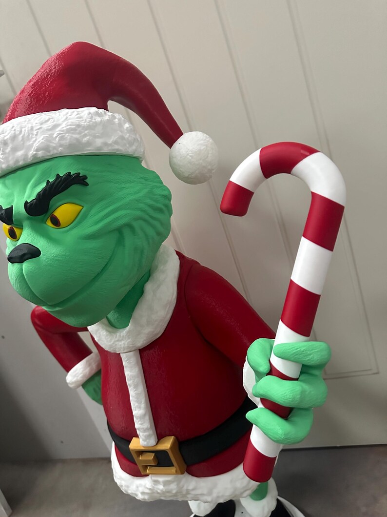 May include: A green character statue wearing a red Santa suit with a black belt and gold buckle. The statue has a red Santa hat with white trim and is holding a red and white striped candy cane. The character has yellow eyes.