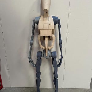 May include: A partially assembled, unpainted, plastic model of a Star Wars battle droid. The droid is standing upright with its arms extended. The droid is made of beige and gray plastic.
