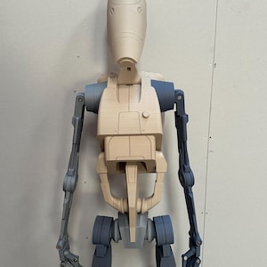 May include: A beige and gray articulated robot figure with a rounded head and jointed limbs. The figure is posed standing with its arms extended.