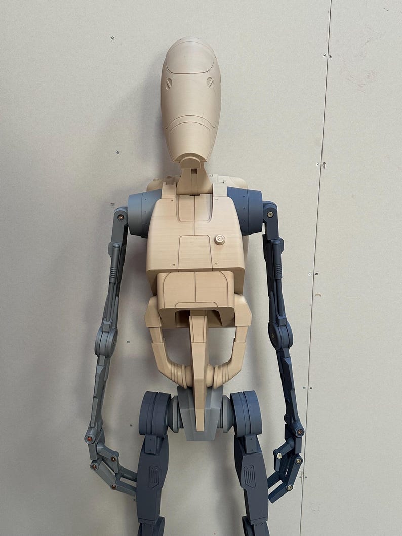 May include: A beige and gray articulated robot figure with a rounded head and jointed limbs. The figure is posed standing with its arms extended.