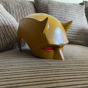 May include: A yellow Daredevil mask with red eyes. The mask is made of plastic and has a textured surface.
