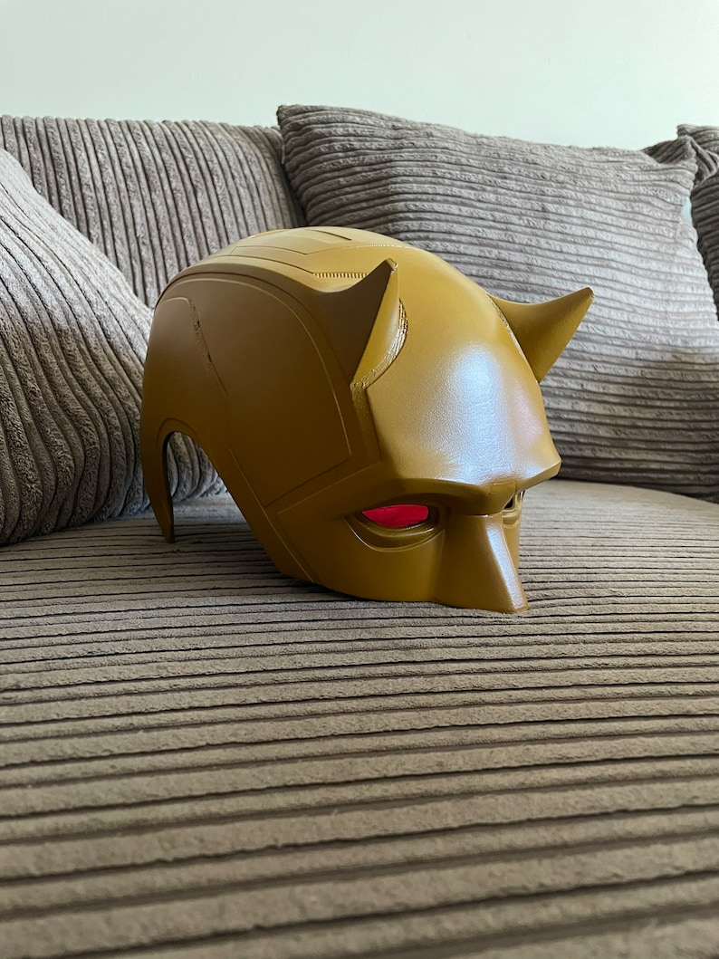 May include: A yellow Daredevil mask with red eyes. The mask is made of plastic and has a textured surface.