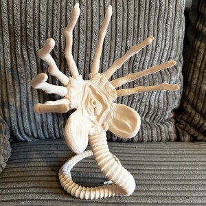Op de afbeelding: Een wit plastic model van een buitenaards wezen met een lange, gesegmenteerde staart en meerdere armen. Het wezen heeft een grote, open mond en een prominent hoofd.