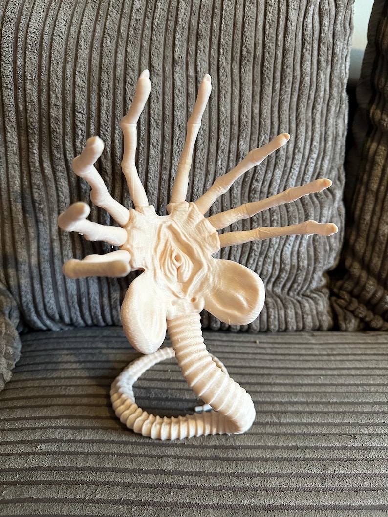 Op de afbeelding: Een wit plastic model van een buitenaards wezen met een lange, gesegmenteerde staart en meerdere armen. Het wezen heeft een grote, open mond en een prominent hoofd.