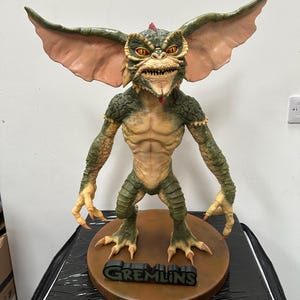 May include: A detailed figurine of a Gremlin, featuring a green and tan color scheme. The creature has large ears, sharp teeth, and a red crest. The figurine stands on a brown base with the word "GREMLINS" in black.