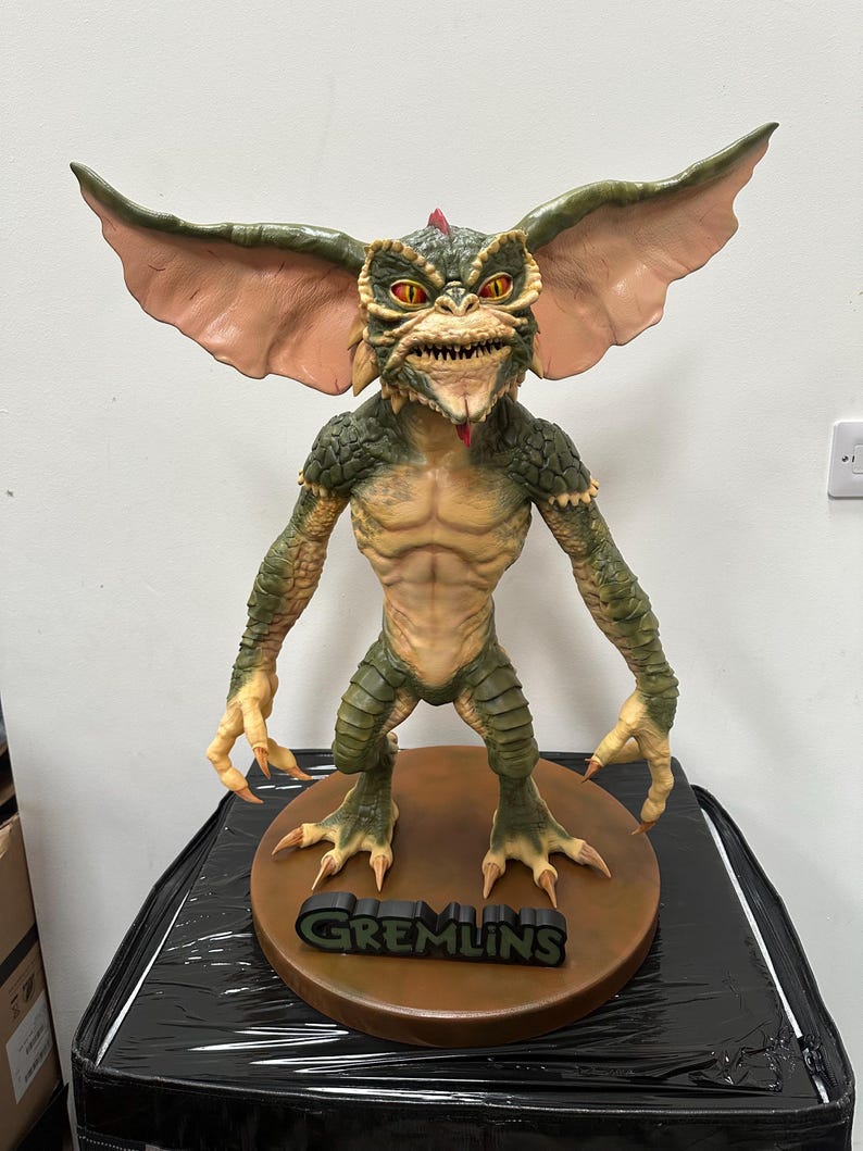 May include: A detailed figurine of a Gremlin, featuring a green and tan color scheme. The creature has large ears, sharp teeth, and a red crest. The figurine stands on a brown base with the word "GREMLINS" in black.