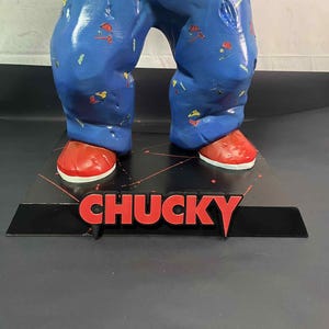 May include: A figurine of a character wearing blue pajamas with colorful patterns and red shoes. The word "CHUCKY" is displayed in red letters on a black base.