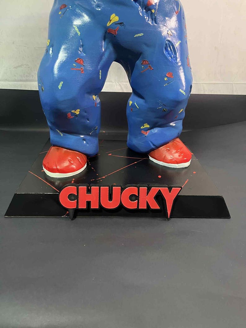 May include: A figurine of a character wearing blue pajamas with colorful patterns and red shoes. The word "CHUCKY" is displayed in red letters on a black base.