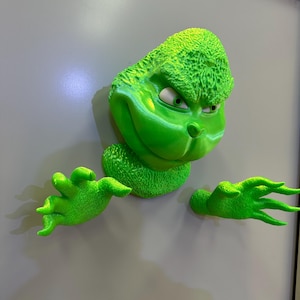 May include: A green Grinch character figurine with a mischievous expression, peeking out from behind a white door. The figurine has textured fur and is holding out its hands.