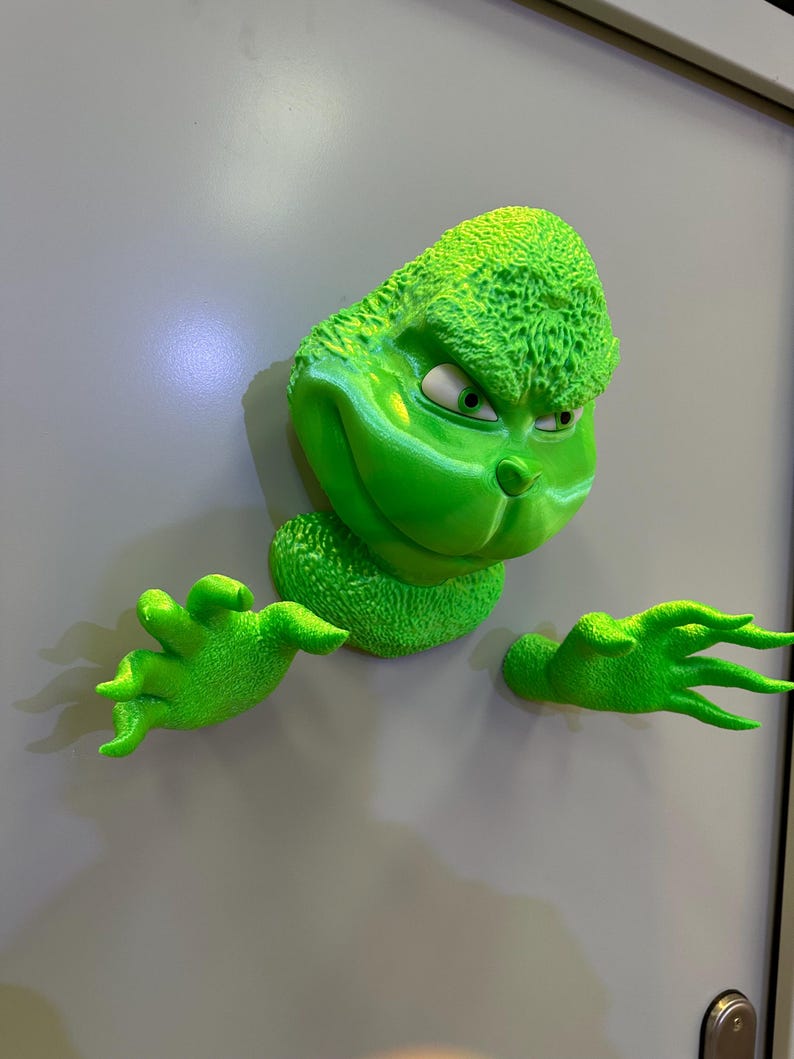 May include: A green Grinch character figurine with a mischievous expression, peeking out from behind a white door. The figurine has textured fur and is holding out its hands.