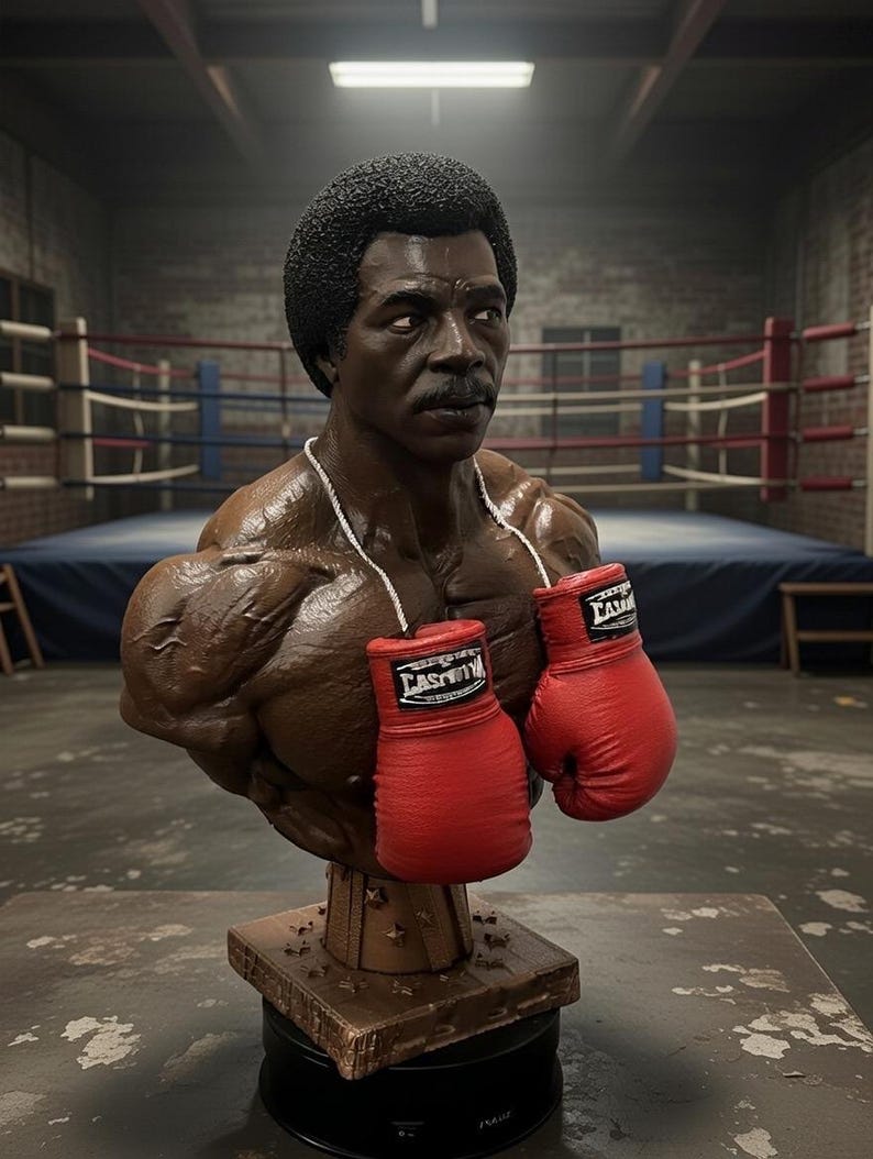 May include: A bust of a muscular man with a dark complexion, mustache, and afro hairstyle. The bust wears a white rope necklace and holds two red boxing gloves. The bust is on a wooden stand, with a boxing ring in the background.