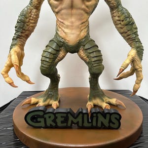 May include: A detailed figurine of a Gremlin creature, featuring a tan torso, green scaled limbs, and sharp claws. The figure stands on a brown base with the word "GREMLINS" in green letters.