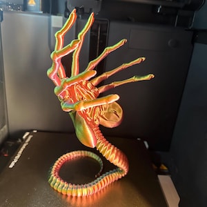 Op de afbeelding: Een 3D-geprinte alienhand met een gesegmenteerde staart, geprint in een regenboogkleurig, iriserend kleurenschema. De hand is opgeheven in een grijpende positie, met wijd gespreide vingers. De staart is om de basis van de hand gekruld.