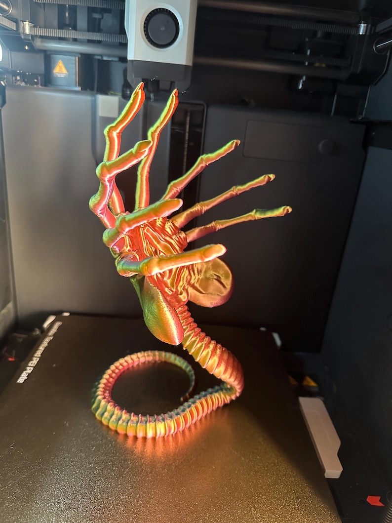 Op de afbeelding: Een 3D-geprinte alienhand met een gesegmenteerde staart, geprint in een regenboogkleurig, iriserend kleurenschema. De hand is opgeheven in een grijpende positie, met wijd gespreide vingers. De staart is om de basis van de hand gekruld.