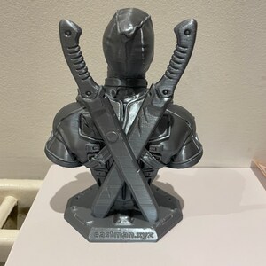 May include: A gray 3D printed bust of Deadpool with his swords crossed behind him. The bust is on a base with the text "eastman.xyz" printed on it.