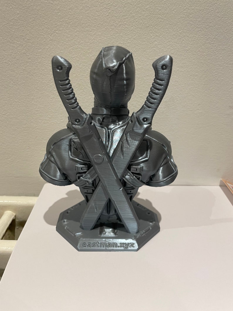 May include: A gray 3D printed bust of Deadpool with his swords crossed behind him. The bust is on a base with the text "eastman.xyz" printed on it.