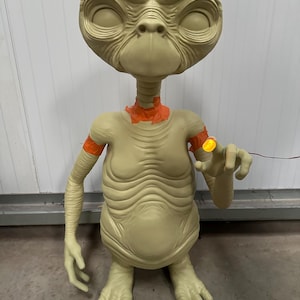 May include: A beige-colored, life-size E.T. the Extra-Terrestrial figure. The figure has large eyes, a long neck, and a wrinkled appearance. It has orange accents on the neck and arms, and a glowing finger. The figure is standing.