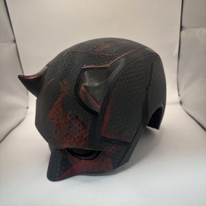 Könnte beinhalten: Ein schwarzer und roter Superheldenhelm mit strukturierter Oberfläche. Der Helm hat spitze Hörner und einen Visierbereich. Die roten Akzente sind über die Oberfläche gespritzt. Der Helm ist wahrscheinlich ein Cosplay- oder Sammlerartikel.