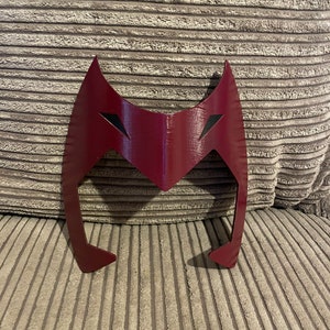 May include: A red plastic mask with a pointed top and two eye holes. The mask is shaped like a stylized head with a pointed chin.