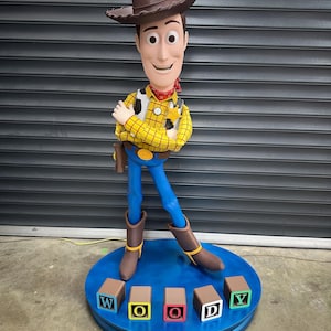 May include: A large statue of the Woody character from Toy Story. Woody is depicted in a brown cowboy hat, yellow plaid shirt, blue trousers, and brown boots. The statue stands on a blue base with blocks spelling "WOODY".