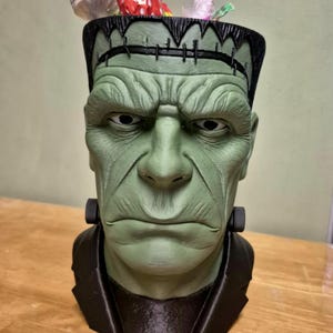 Puede incluir: Un recipiente verde con forma de cabeza de monstruo de Frankenstein con borde y cuello negros. El recipiente est&aacute; lleno de caramelos envueltos. La cara del monstruo tiene rasgos detallados y una expresi&oacute;n severa.