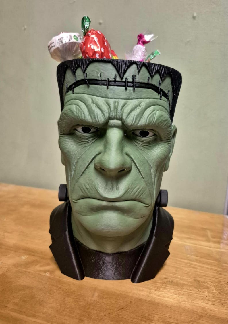 Puede incluir: Un recipiente verde con forma de cabeza de monstruo de Frankenstein con borde y cuello negros. El recipiente est&aacute; lleno de caramelos envueltos. La cara del monstruo tiene rasgos detallados y una expresi&oacute;n severa.